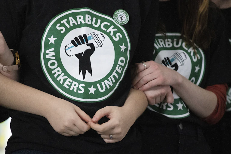 Starbucks employees and supporters link arms, with two wearing Starbucks Workers United t-shirts.
