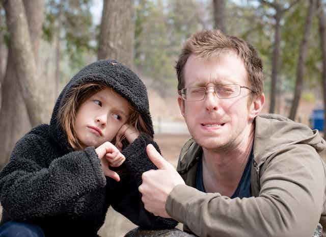 Young man squints, grimaces and makes a thumbs-up gesture while his young, glum-looking daughter looks at him disapprovingly.