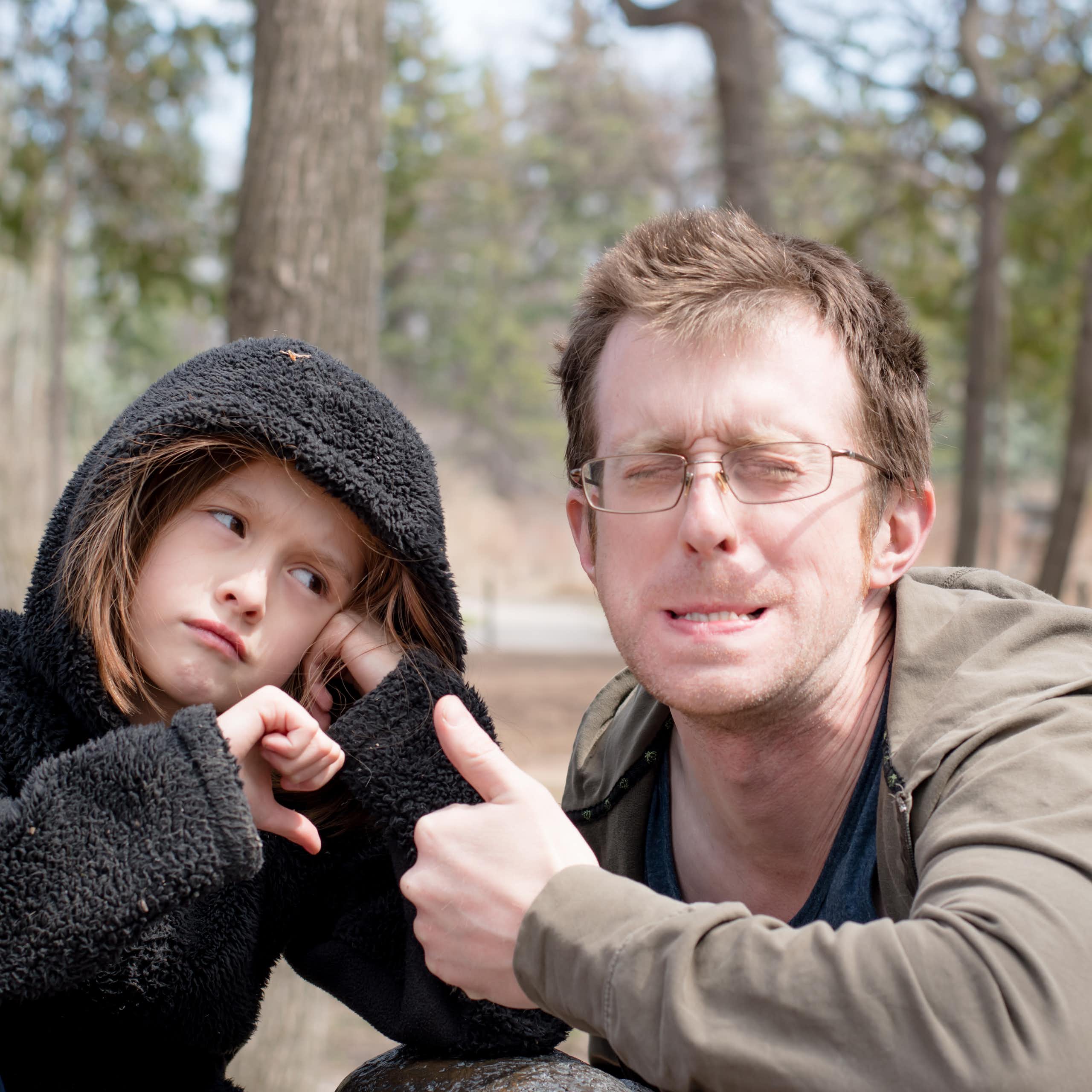 Young man squints, grimaces and makes a thumbs-up gesture while his young, glum-looking daughter looks at him disapprovingly.