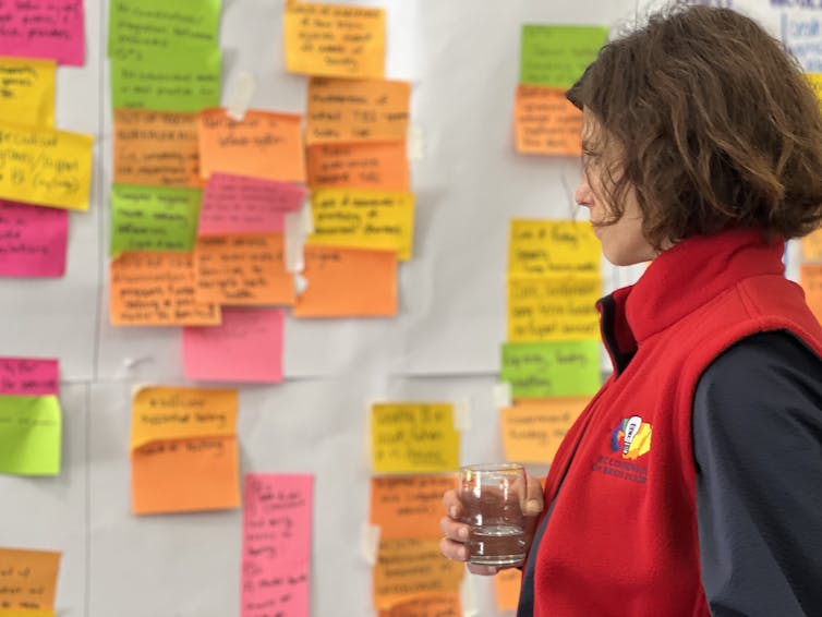 A woman in a red vest looking at a white board covered with Post-It notes.