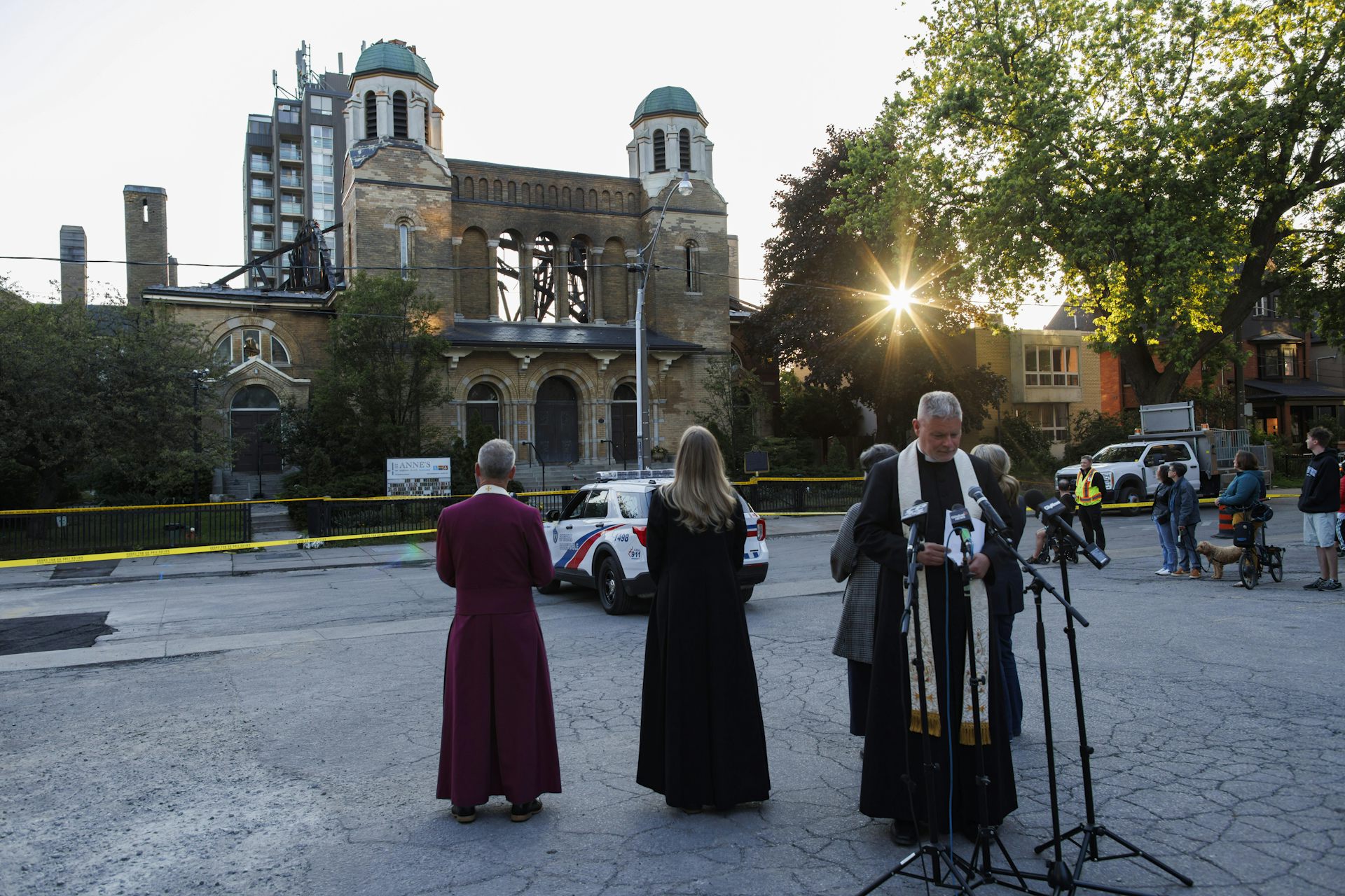A priest in a robe seen leading an outdoor gathering with head bowed in front of a burned out structure.