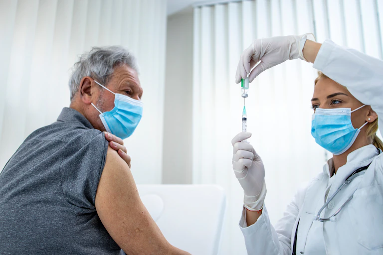 A health-care worker drawing up a vial of vaccine, while a man sits with his sleeve rolled up.