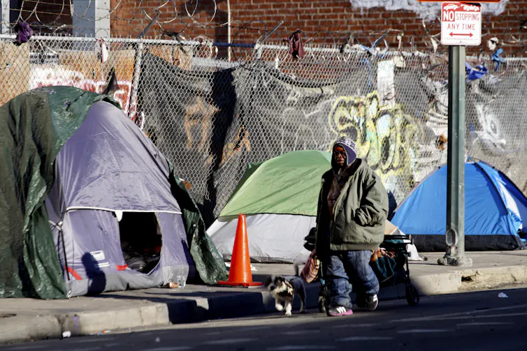 A man walks a dog on a leash past tents on a sidewalk