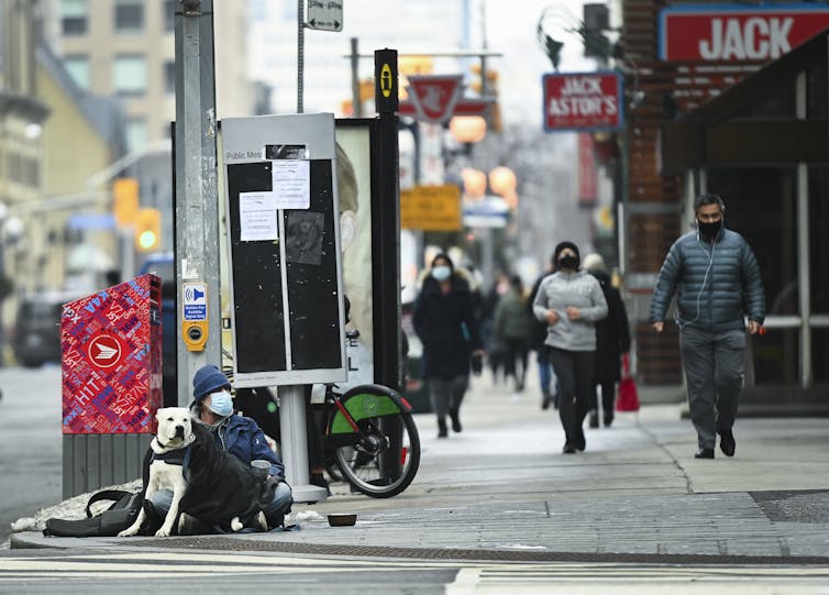 A person in a hoodie and face mask sits on a sidewalk with a white dog. Other people walk past.