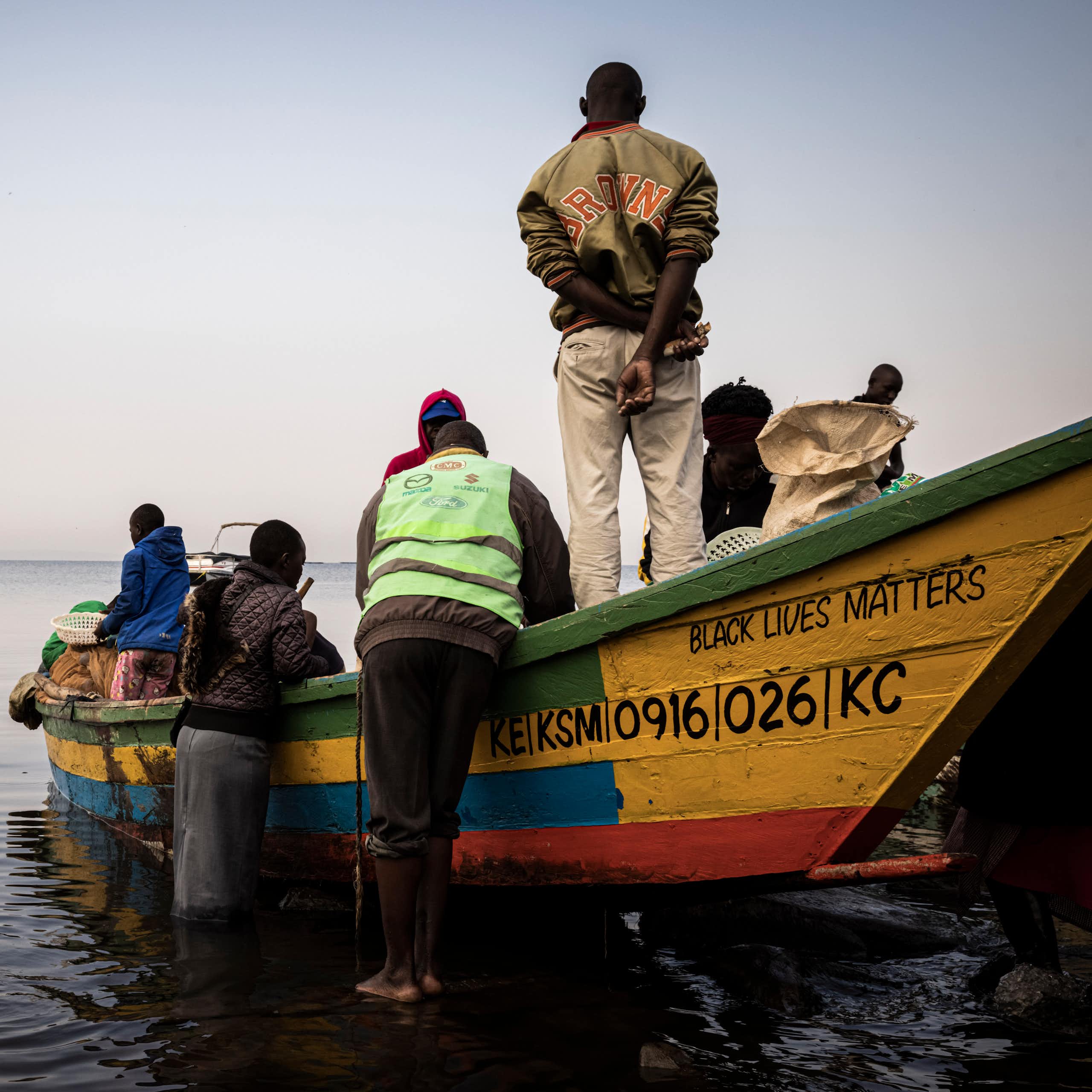 A wooden fishing boat with the words 'Black Lives Matter' on the side and six fisher folk standing in and around it. A woman carries a plastic bucket of fish away from the boat
