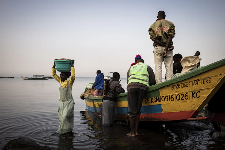 A wooden fishing boat with the words 'Black Lives Matter' on the side and six fisher folk standing in and around it. A woman carries a plastic bucket of fish away from the boat