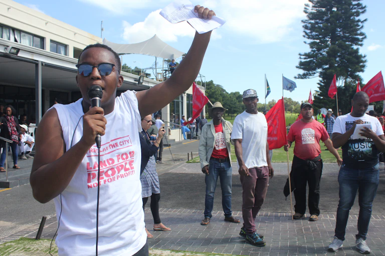 A man talks into a mic and holds his hand up in a fist, behind him people holding flags and banners.
