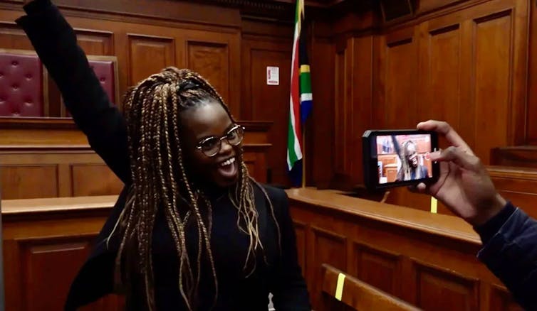 A woman with braids beams as she holds an arm in the air in a wood-panelled courtroom, a hand taking her photo in the foreground.