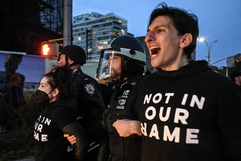 A man wearing a shirt that says 'NOT IN OUR NAME' is pulled away by police.