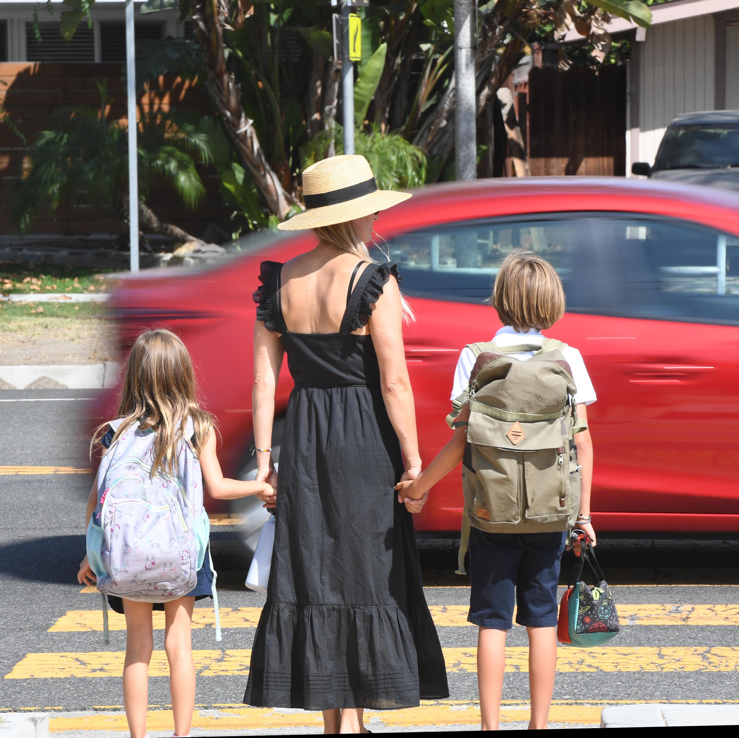 A car speeds past a woman holding two children's hands, standing at a crosswalk