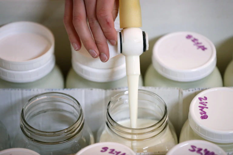 Hand holding nozzle dispensing milk into a glass jar, among an array of other milk-filled glass jars with lids.