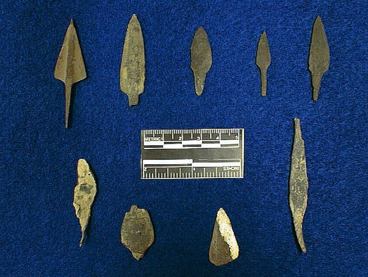 Nine differently shaped copper projectile points rest on a fabric background, with a scale in between.