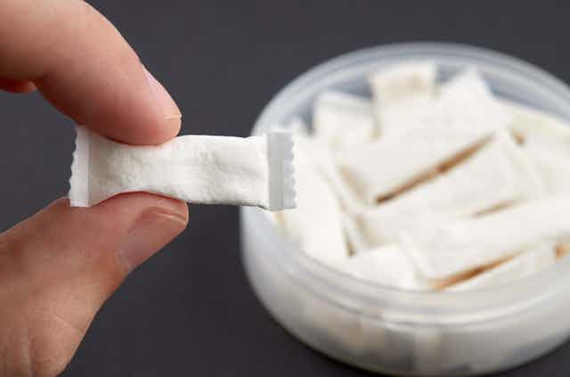Two fingers hold a tiny pouch containing white powder, with a bowl of the same pouches in the background.