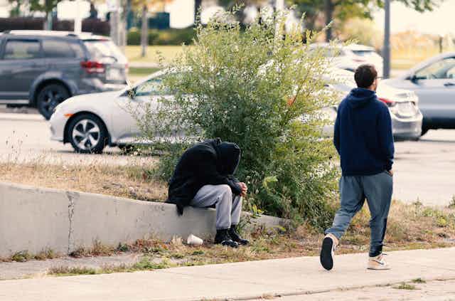 A man walks past a person huddled on a curb