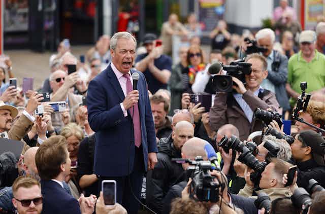 Nigel Farage speaks into a microphone surrounded by a crowd and photographers