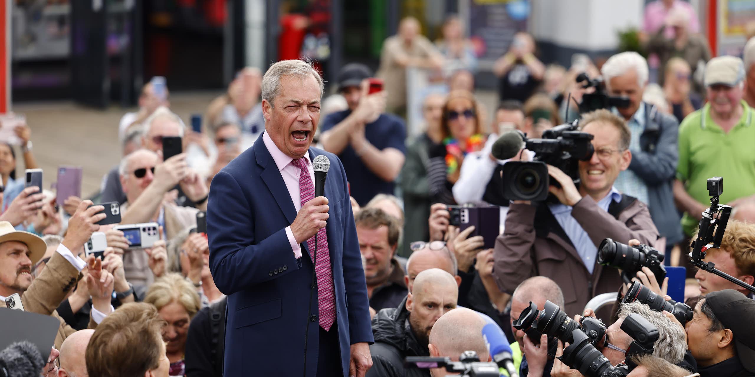 Nigel Farage speaks into a microphone surrounded by a crowd and photographers