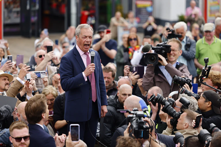 Nigel Farage speaks into a microphone surrounded by a crowd and photographers