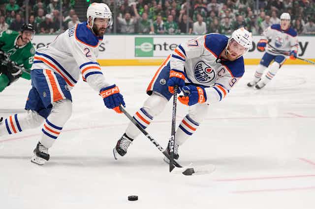 Two men in heavily padded blue, orange and white uniforms skate on ice while holding hockey sticks.