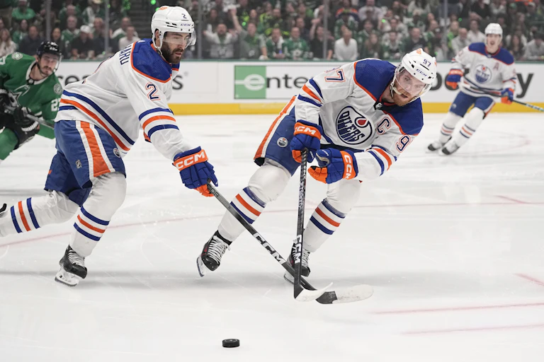 Two men in heavily padded blue, orange and white uniforms skate on ice while holding hockey sticks.