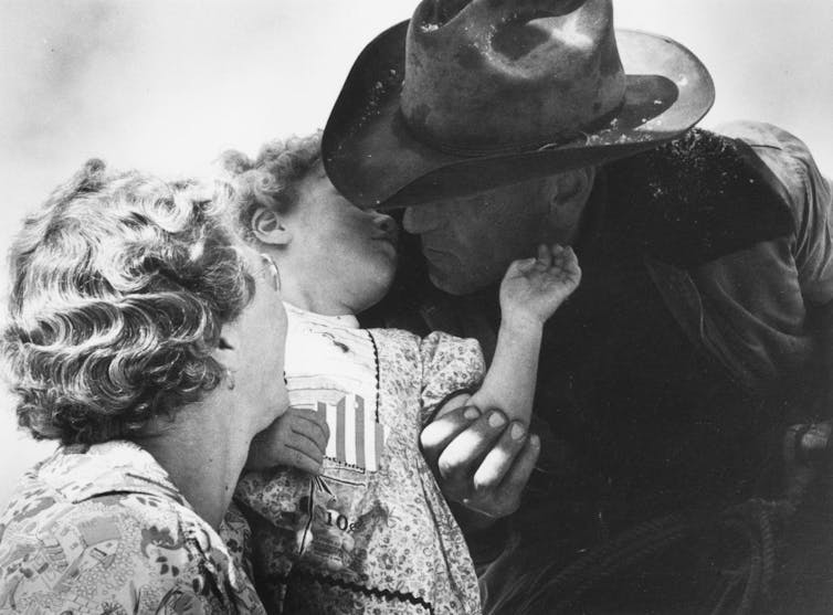 A black and white photograph of a man in a cowboy hat kissing a small child held by a blonde woman.