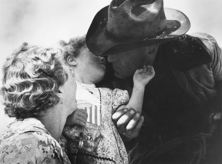 A black-and-white photo of a man in a cowboy hat kissing a small child held by a blonde woman.