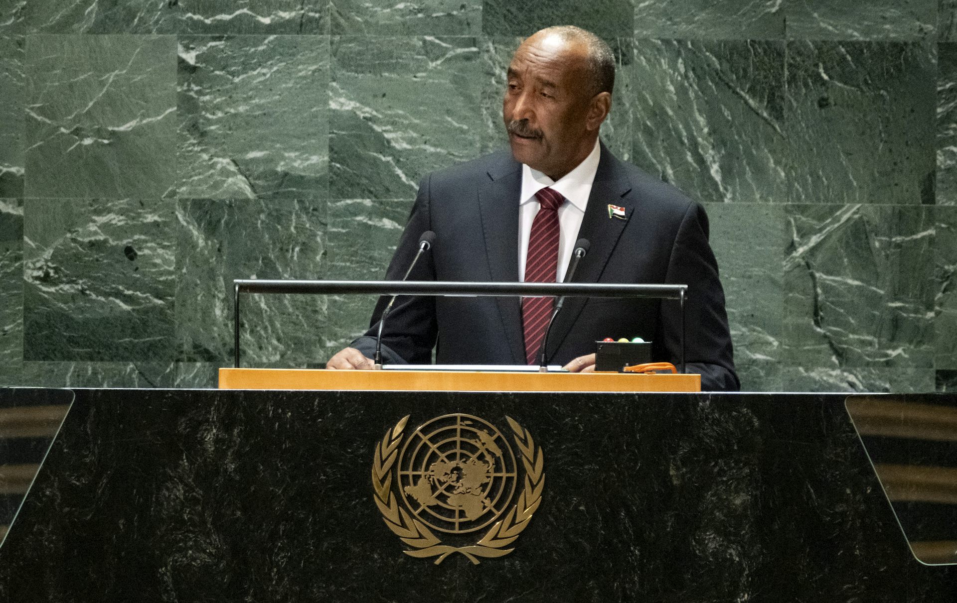 A man in a dark suit stands behind a lectern emblazoned with the United Nations insignia.