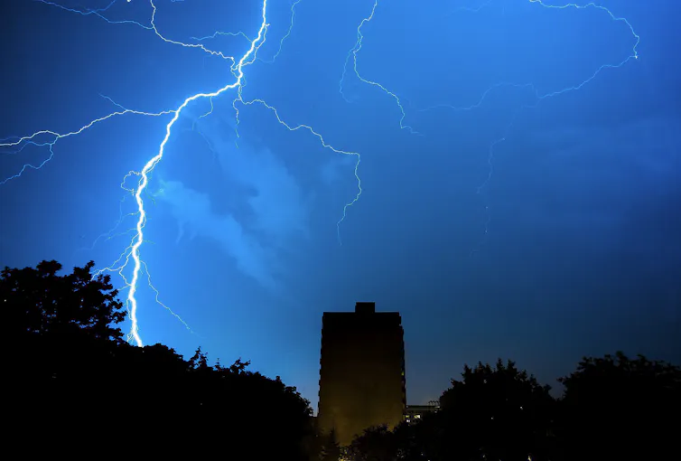 A lightning bolt over trees and an apartment building in silhouette.