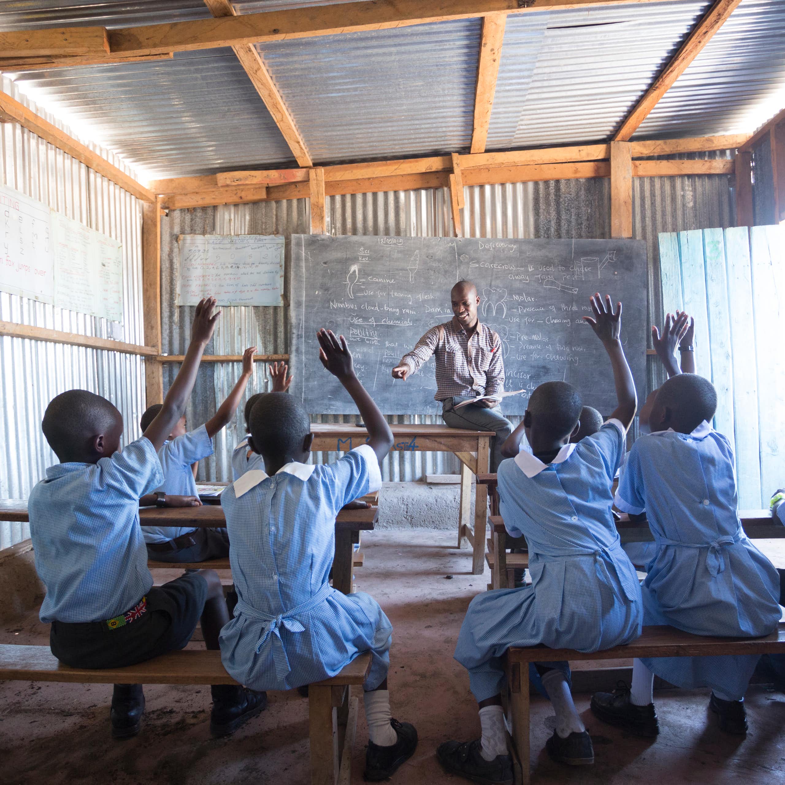 Un pequeño grupo de escolares con uniforme azul claro levantan la mano para dirigirse a un profesor sentado en un pupitre a la cabeza de la clase.