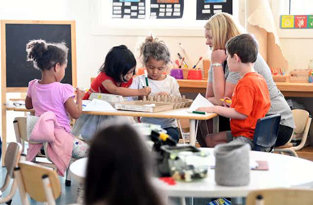 A woman sits at a table with young children in a classroom. There are egg cartons and markers on the table. 