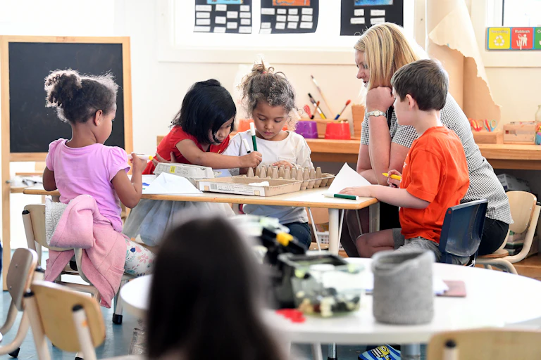A woman sits at a table with young children in a classroom. There are egg cartons and markers on the table.