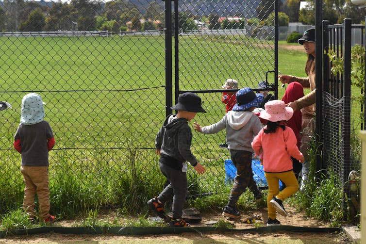 A woman leads a group of children in hats onto onto a large grassy area.