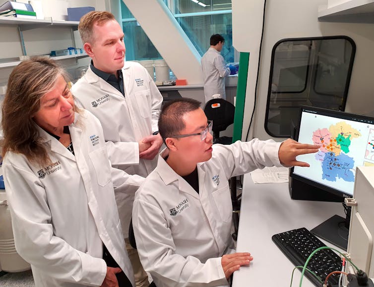 Photo of three people in lab coats looking at a computer screen showing some kind of complex molecular diagram