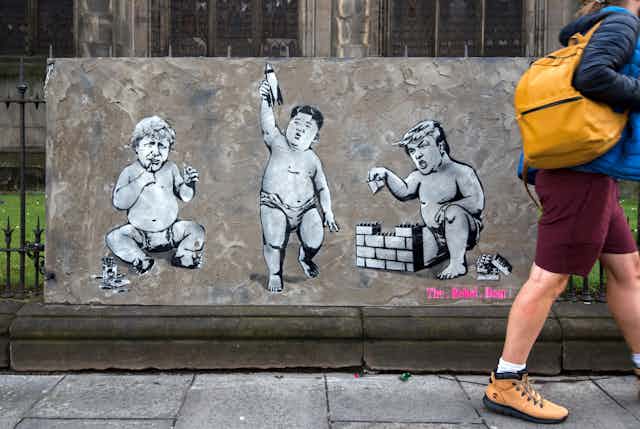 A person walking past street art that depicts Donald Trump, Boris Johnson and Kim Jong un as babies playing with toys.
