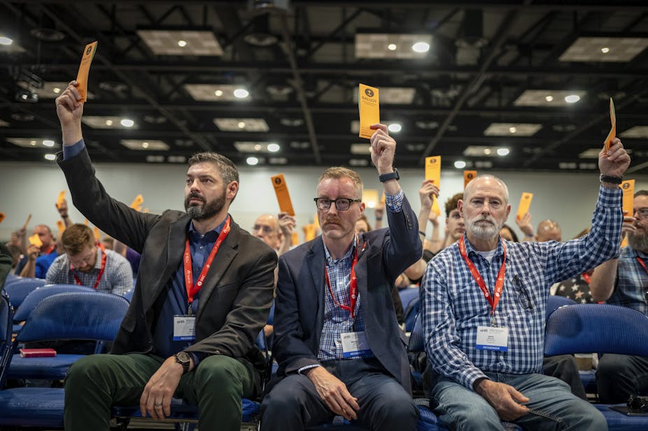 Several people, seated in a large room and wearing name tags, raise their hands while holding yellow cards.