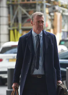 Adam Price wearing a navy jacket and navy tie walking down the street.
