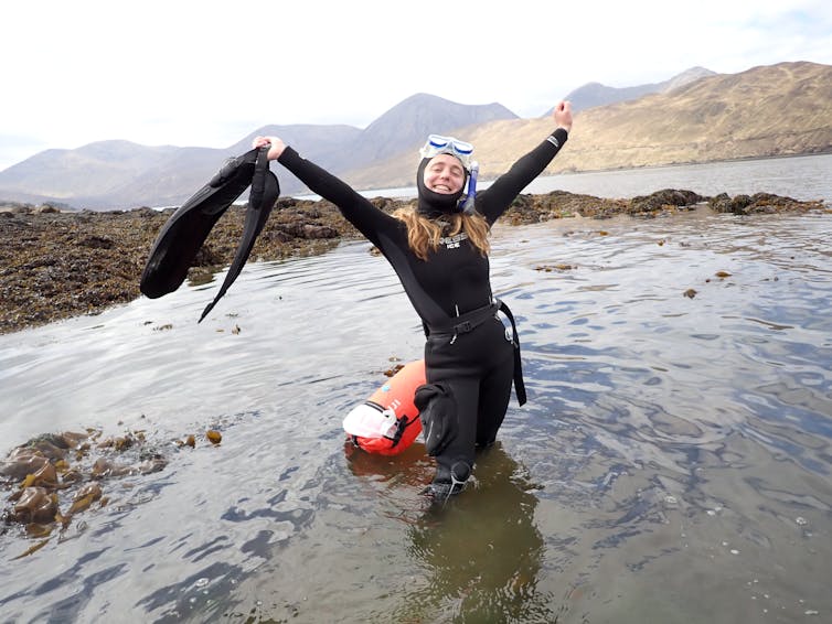 Woman in black wetsuit and snorkel mask, standing in shallow water with orange tow float, arms open wide holding finds, rocky shore behind