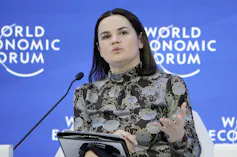 A woman with dark hair wearing a grey and black flowered top speaks into a microphone. A blue World Economic Forum banner is behind her.