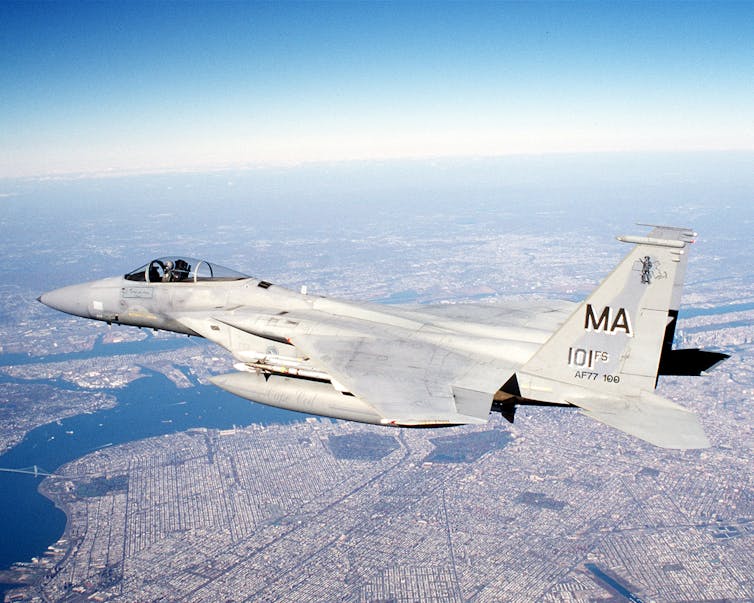 A small fighter airplane is seen in the sky, over a city and rivers below with a clear blue sky behind it.