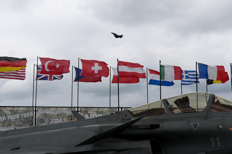 Two rows of country flags, including that of France, Germany and Italy, are shown flying against a grey sky. A large fighter airplane is in front of the flags and also flying in the sky.