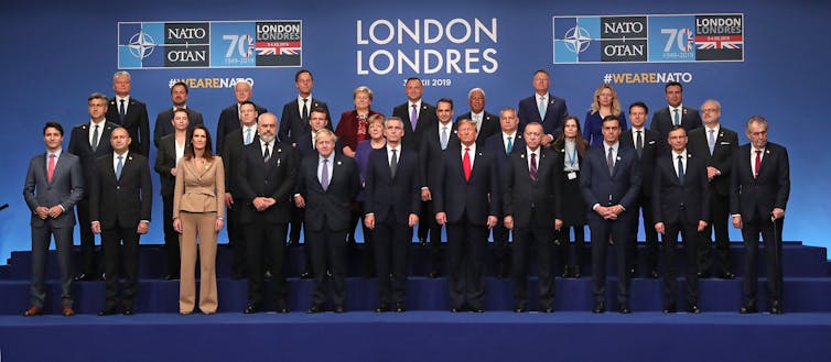 Three rows of people stand on a blue platform, behind which are the words “NATO” and “London”.