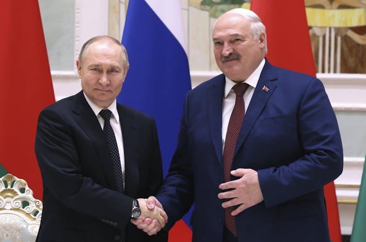 Two balding men in dark suits, one about six inches taller than the other, shake hands with red and blue flags behind them.