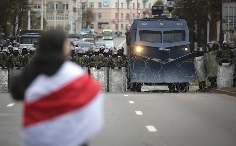 A person seen from behind is draped in a red and white flag as a line of armored police and vehicles approach her down a city street.