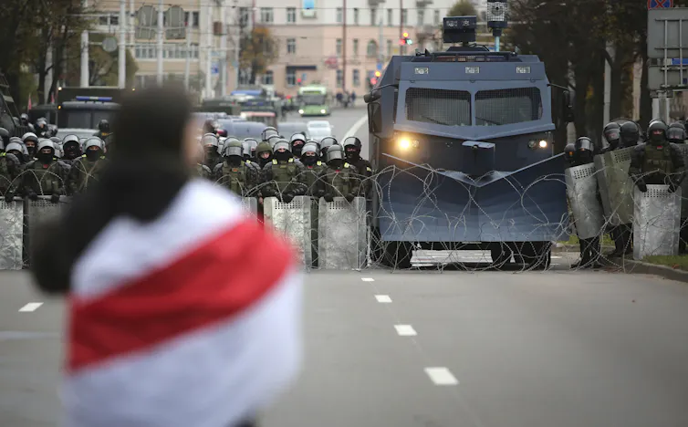 A person seen from behind is draped in a red and white flag as a line of armored police and vehicles approach her down a city street.