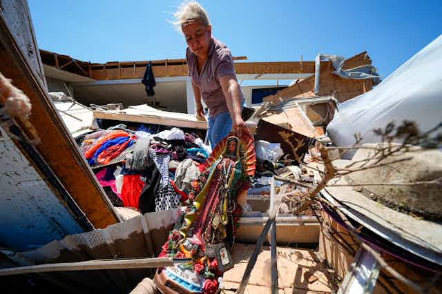 A woman pulls a religious statue out from among debris. A splintered home and scattered clothing are around her.