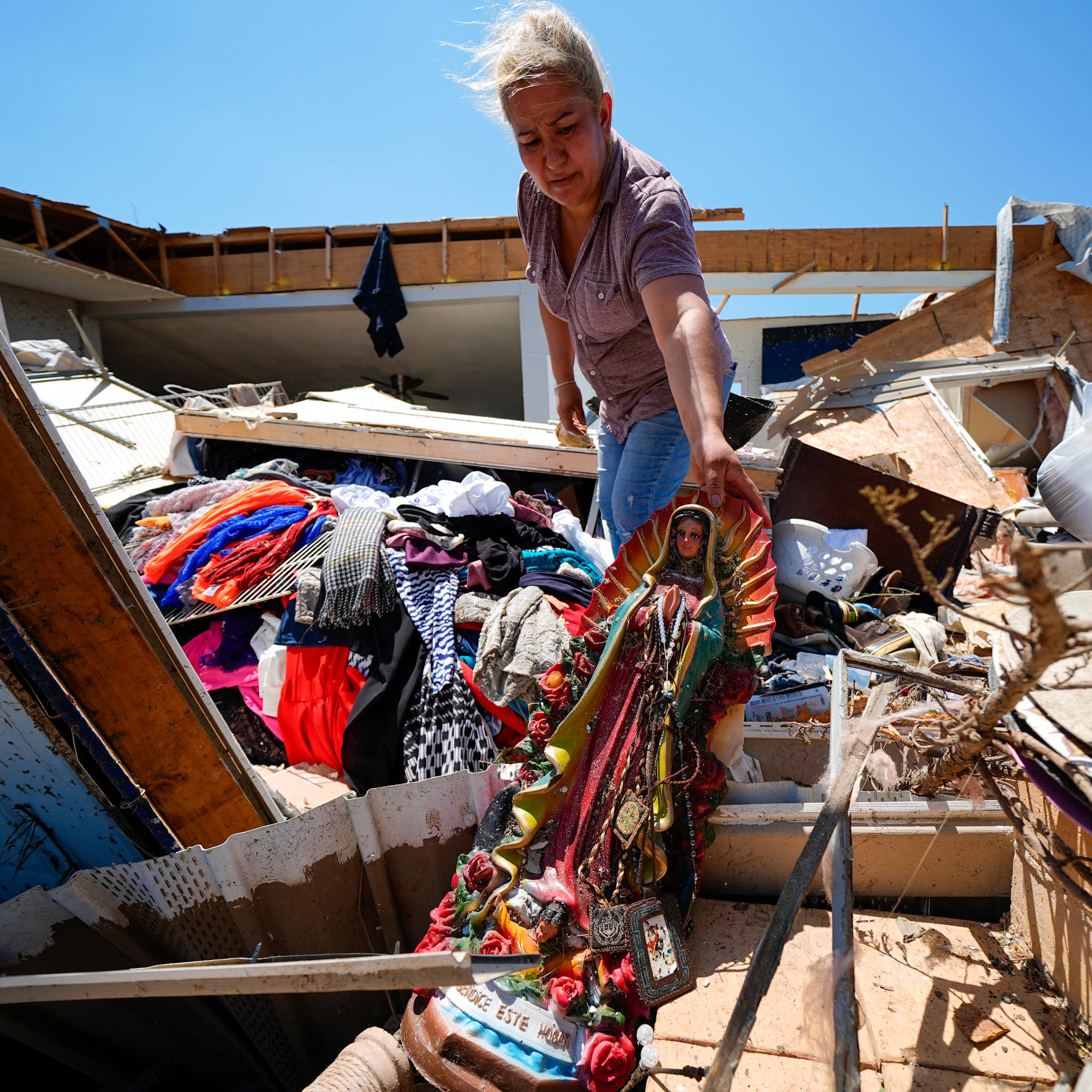 A woman pulls a religious statue out from among debris. A splintered home and scattered clothing are around her.