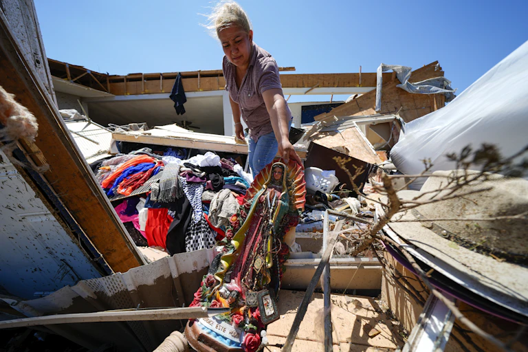 A woman pulls a religious statue out from among debris. A splintered home and scattered clothing are around her.