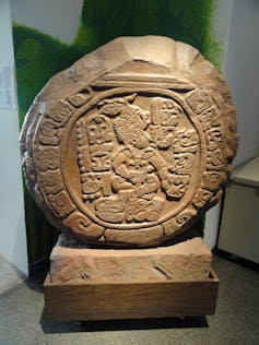 An earthen-colored circle with inscriptions and a human figure in the middle rests on top of a short pedestal.