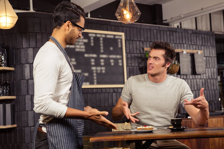 A man pointing at a cup of coffee and a cake, looking angrily at a waiter.