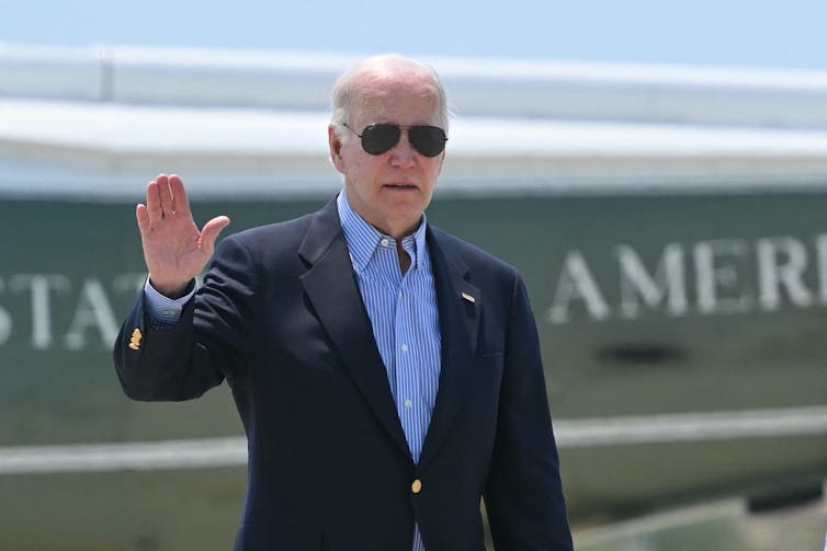 A white man with white hair and a dark suit and sunglasses holds up his hand to the camera.