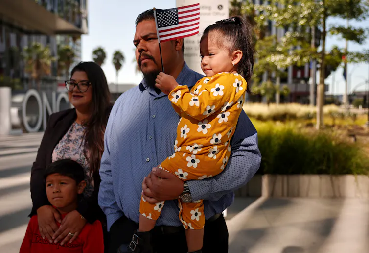 A man holds a small child, who holds an American flag. He stands next to a woman, who places her hands around the chest of a young boy.